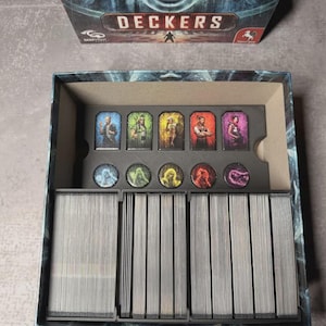 May include: An open board game box labelled "DECKERS". Inside are character cards and game pieces. The cards display character illustrations. The game pieces are round tokens. The box sits on a grey surface.