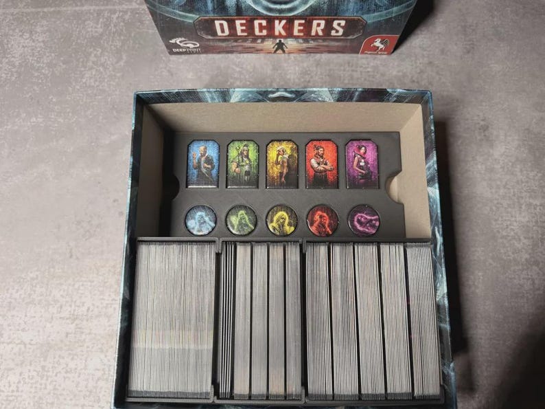 May include: An open board game box labelled "DECKERS". Inside are character cards and game pieces. The cards display character illustrations. The game pieces are round tokens. The box sits on a grey surface.