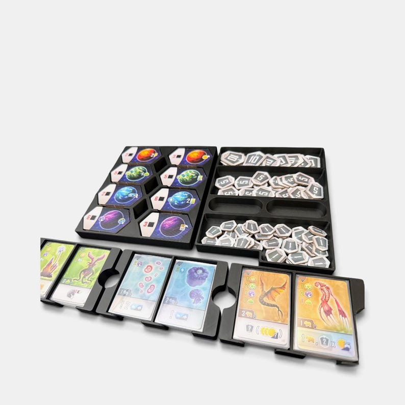 K&ouml;nnte beinhalten: Ein schwarzes Kunststoff-Organizer-Set f&uuml;r Brettspiele mit sechseckigen Planetentafeln, kleinen wei&szlig;en sechseckigen Zahlenmarkern und mehreren Stapeln rechteckiger Spielkarten, die bunte Illustrationen von Kreaturen und verschiedene Spielsymbole zeigen.