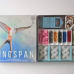 Op de afbeelding: Open Wingspan bordspeldoos. De doosafbeelding toont een vogel met uitgespreide vleugels. Binnenin zitten spelstukken in verschillende kleuren, waaronder roze, bruin, groen, blauw en rood. Er zijn ook kaarten en een blauwe boot.