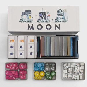 Può includere: Gioco da tavolo "MOON" con illustrazioni di veicoli spaziali. La scatola contiene pile di carte, pedine bianche e gettoni colorati in vassoi. I componenti del gioco sono ben organizzati.
