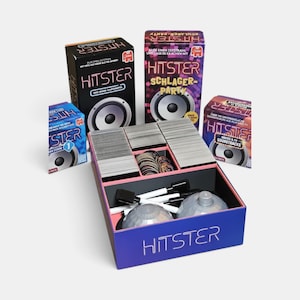Può includere: Un set di gioco di carte Hitster con una grande scatola blu contenente mazzi di carte, gettoni circolari, pennarelli bianchi e due oggetti traslucidi a forma di cupola. Tre scatole di gioco Hitster aggiuntive sono esposte dietro.