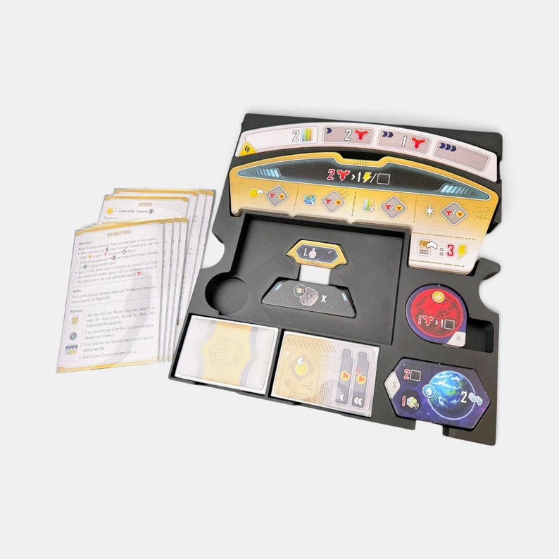 K&ouml;nnte beinhalten: Ein schwarzes Kunststoff-Organizer-Tablett f&uuml;r Brettspiele mit verschiedenen Spielkomponenten, darunter ein Stapel wei&szlig;er Anleitungskarten, ein gelb-schwarzes Dashboard, ein runder roter Spielstein und ein sechseckiges blaues Planetenpl&auml;ttchen mit der Zahl 2.