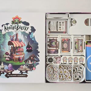 Peut inclure: Boîte de jeu de société ouverte avec le titre "Apothecary" et le sous-titre "The Culinary Alchemists". La boîte contient des cartes de jeu, des jetons et un récipient bleu avec un logo blanc. L'illustration de la boîte présente une illustration fantaisiste d'une boulangerie.