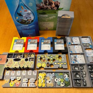 Puede incluir: Un organizador de juego para el juego de mesa Ark Nova. El organizador está hecho de plástico gris y tiene compartimentos para todos los componentes del juego, incluidas las cartas, las fichas y las losetas. El organizador está diseñado para ayudar a los jugadores a mantener su juego organizado y ordenado.