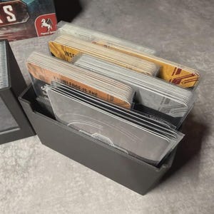May include: A grey card holder filled with stacks of cards. Some cards have text that reads "KNOWLEDGE IS THE". The cards are in clear plastic sleeves. A board game box is visible in the background.