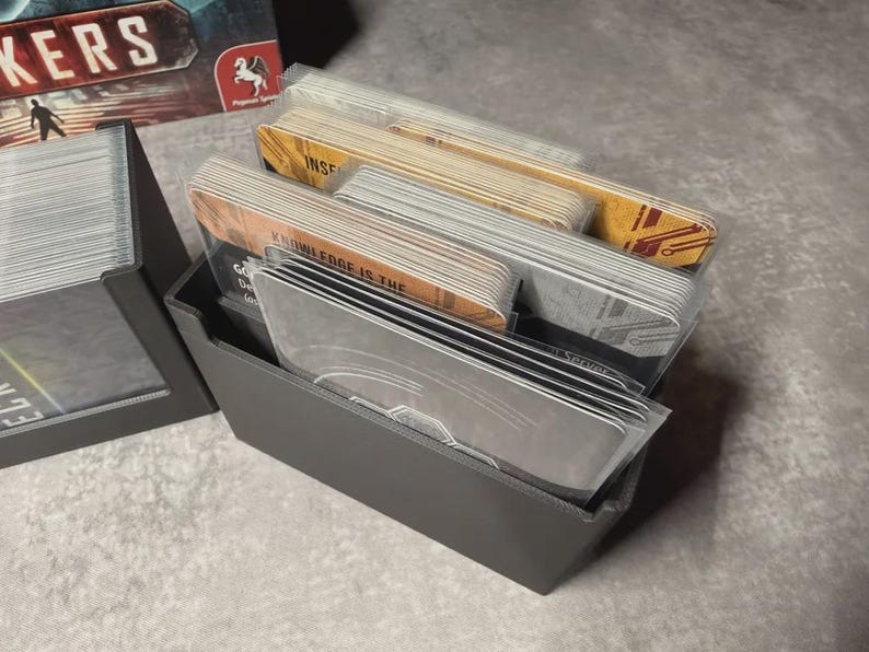 May include: A grey card holder filled with stacks of cards. Some cards have text that reads "KNOWLEDGE IS THE". The cards are in clear plastic sleeves. A board game box is visible in the background.