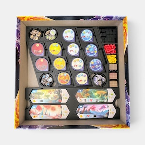K&ouml;nnte beinhalten: Ein Brettspiel-Box-Einsatz mit organisierten Spielkomponenten. Das schwarze Kunststoff-Inlay enth&auml;lt runde Spielsteine mit bunten Symbolen, Stapel aus kleinen schwarzen, roten, gelben und beigen W&uuml;rfeln sowie vier rechteckige Spielertableaus mit Landschaftsbildern.