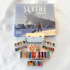 Puede incluir: El juego de mesa Scythe Legendary Box se sitúa sobre organizadores de plástico blanco llenos de varios componentes de juego, incluyendo pequeñas fichas de madera de colores, marcadores de recursos circulares y mazos de cartas.