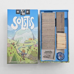 Könnte beinhalten: Eine Brettspielbox und ihr Inhalt. Die Box ist hellblau mit dem Wort "Solstis" in Weiß. Das Box-Artwork zeigt eine Cartoon-Landschaft mit einem Berg und einer Figur. Im Inneren befinden sich Holzspielsteine.