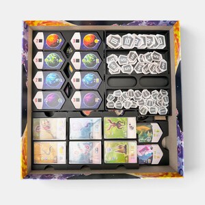 K&ouml;nnte beinhalten: Ein Brettspiel-Box-Einsatz mit organisierten Spielkomponenten. Die linke Seite enth&auml;lt acht sechseckige Planetentafeln mit verschiedenen Farben und Symbolen. Die rechte Seite enth&auml;lt Stapel wei&szlig;er sechseckiger Spielsteine mit Zahlen, und unten befinden sich Reihen rechteckiger Spielkarten mit farbenfrohen Illustrationen.