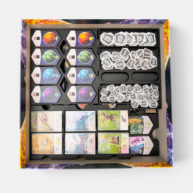 K&ouml;nnte beinhalten: Ein Brettspiel-Box-Einsatz mit organisierten Spielkomponenten. Die linke Seite enth&auml;lt acht sechseckige Planetentafeln mit verschiedenen Farben und Symbolen. Die rechte Seite enth&auml;lt Stapel wei&szlig;er sechseckiger Spielsteine mit Zahlen, und unten befinden sich Reihen rechteckiger Spielkarten mit farbenfrohen Illustrationen.