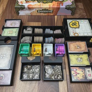 Puede incluir: Un organizador de juegos de mesa con múltiples compartimentos y bandejas. Incluye tarjeteros, organizadores de fichas y un tablero de juego. La caja está etiquetada "EMBERHEART" y presenta un diseño decorativo. La combinación de colores incluye negro, verde, amarillo y morado.