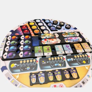 K&ouml;nnte beinhalten: Ein kreisf&ouml;rmiges Brettspiel-Set mit einem zentralen, weltraumthematischen Spielplan und schwarzen Kunststoff-Organizern f&uuml;r Karten und Spielsteine. Das Set enth&auml;lt bunte Planetentafeln, illustrierte Kreaturenkarten und hexagonale Marker.
