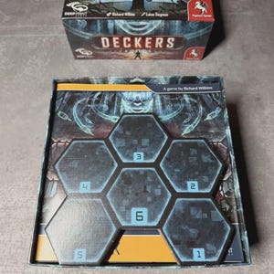 May include: A board game box and game board with a dark, futuristic theme. The box reads "DECKERS" and the game board features hexagonal tiles numbered 1 to 6. The game is by Richard Wilkins.
