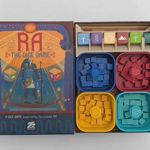 Pode incluir: Caixa do jogo de tabuleiro Ra The Dice Game com uma ilustração de temática egípcia apresentando uma divindade, dois dados e o título. A caixa aberta contém uma bandeja de plástico com seis dados coloridos e quatro tigelas quadradas com cubos de madeira.