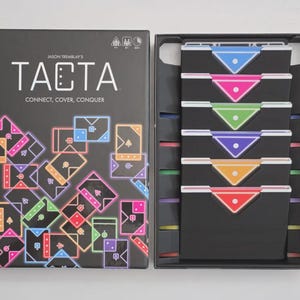 May include: A board game called "TACTA" is shown, with its box and contents. The black box features the game's title and the tagline "CONNECT, COVER, CONQUER." Inside, there are colourful triangular pieces and game components. The game is designed for multiple players.