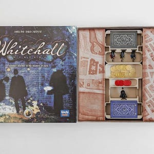 May include: Open board game box titled "Whitehall" with a dark blue and purple cover featuring silhouettes. Inside, compartments hold game pieces, including tokens, figurines, and cards. The game's theme appears to be mystery and investigation.