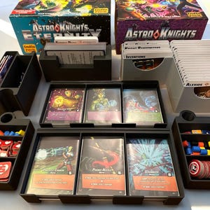 Könnte beinhalten: Ein schwarz-weißer Kartenspiel-Organizer mit mehreren Fächern zum Verstauen von Karten, Spielsteinen und Würfeln. Der Organizer ist mit "Astro Knights Eternity" beschriftet.