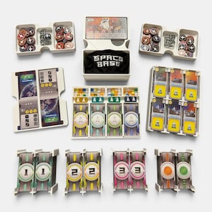 May include: A board game set featuring white plastic organizers containing various game components including circular tokens, dice, small colored cubes, card decks, and player boards. A black fabric bag with the white text SPACE BASE is included.