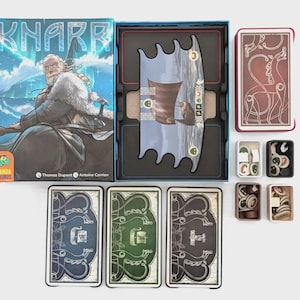Könnte beinhalten: Das Brettspiel "Knarr" wird gezeigt, mit einer Box, die eine Wikinger-Illustration und den Spieltitel zeigt. Das Bild zeigt Spielkomponenten, darunter Karten, Spielsteine und ein Spielbrett, die alle auf einer weißen Oberfläche angeordnet sind.