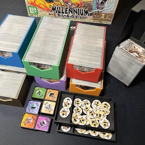 Könnte beinhalten: Eine Sammlung von Millennium Blades-Kartenspielkomponenten, darunter verschiedenfarbige Kartenaufbewahrungsboxen, Spielmarken und Karten. Die Spielbox ist im Hintergrund sichtbar.