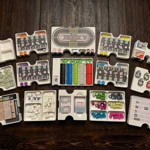 May include: A white plastic game organizer with multiple compartments for storing game pieces, cards, and other components. The organizer has a variety of compartments, including a large central compartment for the game board, smaller compartments for dice, tokens, and cards, and a compartment for the rule book.