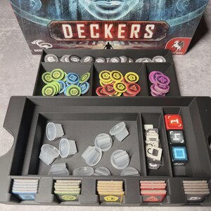 May include: A black game organiser with compartments filled with game pieces. The pieces include coloured tokens, dice, clear plastic containers, and cards. The game box, labelled "DECKERS," is visible in the background.