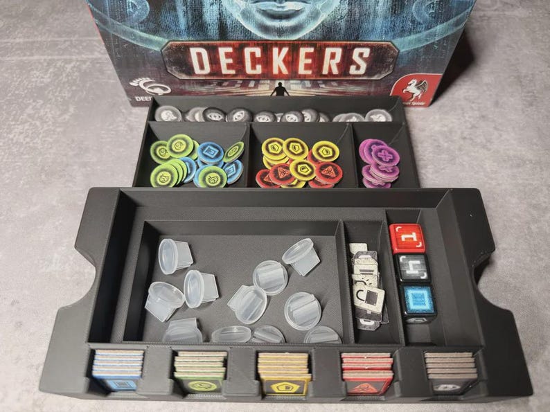 May include: A black game organiser with compartments filled with game pieces. The pieces include coloured tokens, dice, clear plastic containers, and cards. The game box, labelled "DECKERS," is visible in the background.