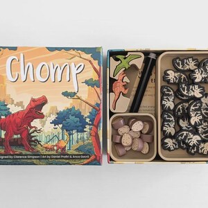 Op de afbeelding: Een bordspel genaamd "Chomp" wordt getoond, met een doosdeksel met een rode dinosaurus. Binnenin de doos bevinden zich verschillende spelonderdelen, waaronder houten dinosaurusspeelstukken, fiches en andere elementen. Het ontwerp bevat een stadsgezicht en bomen.