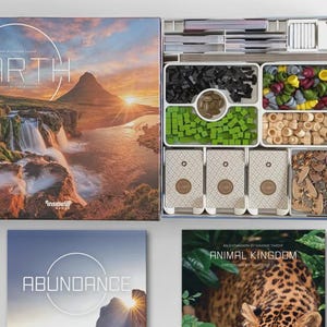 Könnte beinhalten: Geöffnete Brettspielbox mit Komponenten. Der Deckel der Box zeigt eine Landschaft mit Berg, Wasserfall und Sonnenuntergang. Im Inneren befinden sich Spielsteine in weißen Tabletts, darunter Holzmarker, grüne Würfel und schwarze Formen. Zusätzliche Spielbox und Erweiterungsbox sichtbar.
