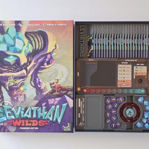 May include: A board game box, open to reveal the contents. The box lid displays the title "Leviathan Wilds" and a fantasy illustration. Inside, the game components are neatly arranged in trays, including cards and game pieces.