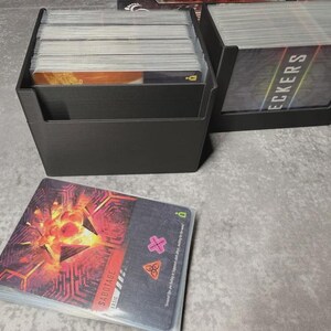 May include: Black card holders filled with playing cards. One holder is in the foreground with a stack of cards in front of it. The cards have a red and orange design with the word "SABOTAGE" on them. The other holder has the word "DECKERS" on the cards.