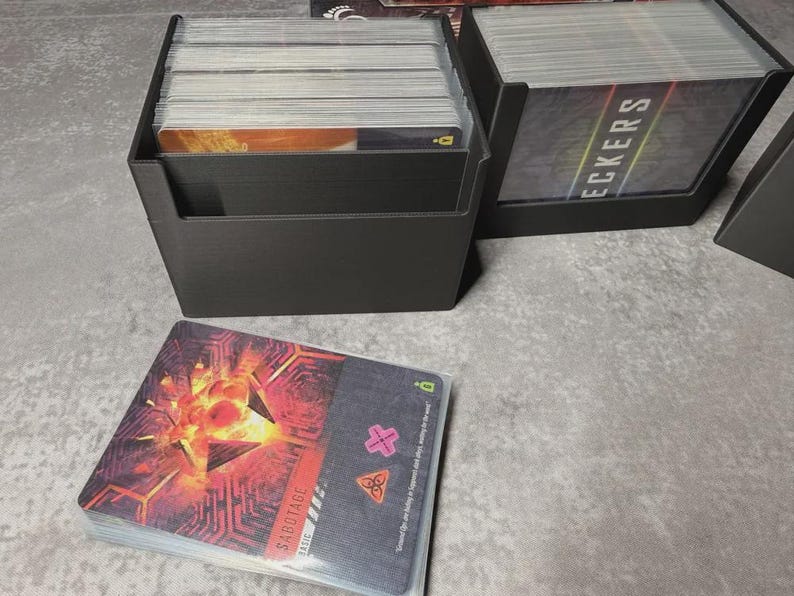 May include: Black card holders filled with playing cards. One holder is in the foreground with a stack of cards in front of it. The cards have a red and orange design with the word "SABOTAGE" on them. The other holder has the word "DECKERS" on the cards.
