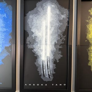 K&ouml;nnte beinhalten: Gerahmtes Kunstwerk mit zwei wei&szlig;en Lichtschwertern vor einem grau-wei&szlig;en Hintergrund. Das Kunstwerk ist vor einem schwarzen Hintergrund mit dem Namen "AHSOKA TANO" unten abgebildet. Der Rahmen ist schwarz.