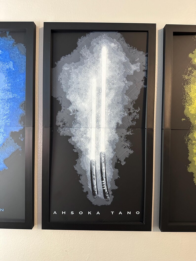 K&ouml;nnte beinhalten: Gerahmtes Kunstwerk mit zwei wei&szlig;en Lichtschwertern vor einem grau-wei&szlig;en Hintergrund. Das Kunstwerk ist vor einem schwarzen Hintergrund mit dem Namen "AHSOKA TANO" unten abgebildet. Der Rahmen ist schwarz.