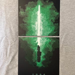 K&ouml;nnte beinhalten: Zwei schwarze Kunstdrucke mit einem gr&uuml;nen Lichtschwert-Design. Einer zeigt den Lichtstrahl, der andere den Griff, mit dem Wort "YODA" unten. Der Hintergrund hat einen gr&uuml;nen, rauchigen Effekt. F&uuml;r Star Wars-Fans.