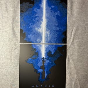 K&ouml;nnte beinhalten: Zweiteiliges Kunstwerk mit einem blau-wei&szlig;en Lichtschwert-Design. Das Lichtschwert hat eine leuchtend wei&szlig;e Klinge vor einem blau-schwarzen Hintergrund. Der Name "ANAKIN" ist unten in Wei&szlig; aufgedruckt.