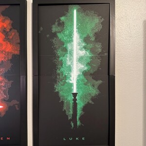 K&ouml;nnte beinhalten: Gerahmter Kunstdruck mit einem gr&uuml;nen Lichtschwert-Design, einer wei&szlig;en Klinge und gr&uuml;nen Energieausbr&uuml;chen auf schwarzem Hintergrund. Der Name "LUKE" ist unten in Wei&szlig; gedruckt. Das Kunstwerk befindet sich in einem schwarzen Rahmen.