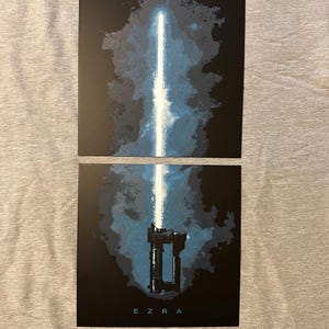 K&ouml;nnte beinhalten: Zwei schwarze Drucke mit einem blauen Lichtschwert-Design. Der obere Druck zeigt die Klinge, der untere Druck den Griff mit dem Namen "EZRA" darunter. Der Hintergrund hat ein blaues und schwarzes abstraktes Design.