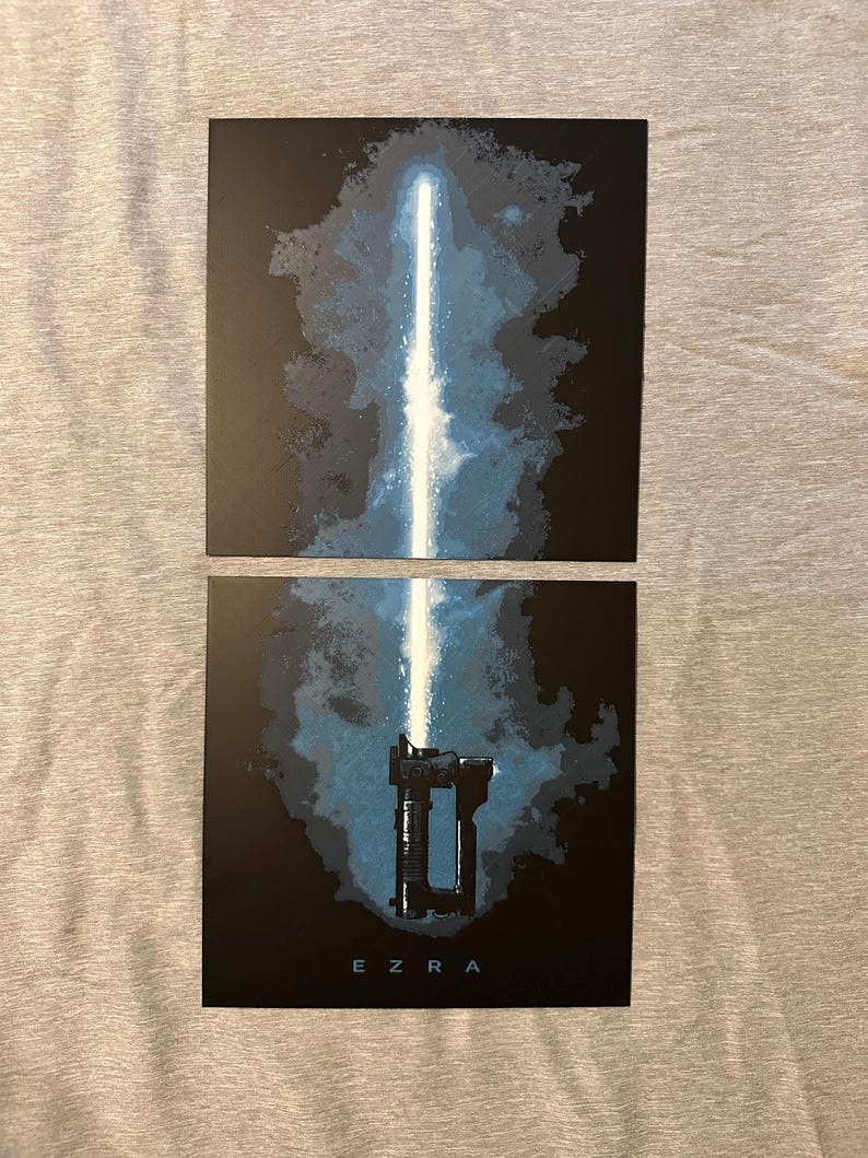 K&ouml;nnte beinhalten: Zwei schwarze Drucke mit einem blauen Lichtschwert-Design. Der obere Druck zeigt die Klinge, der untere Druck den Griff mit dem Namen "EZRA" darunter. Der Hintergrund hat ein blaues und schwarzes abstraktes Design.