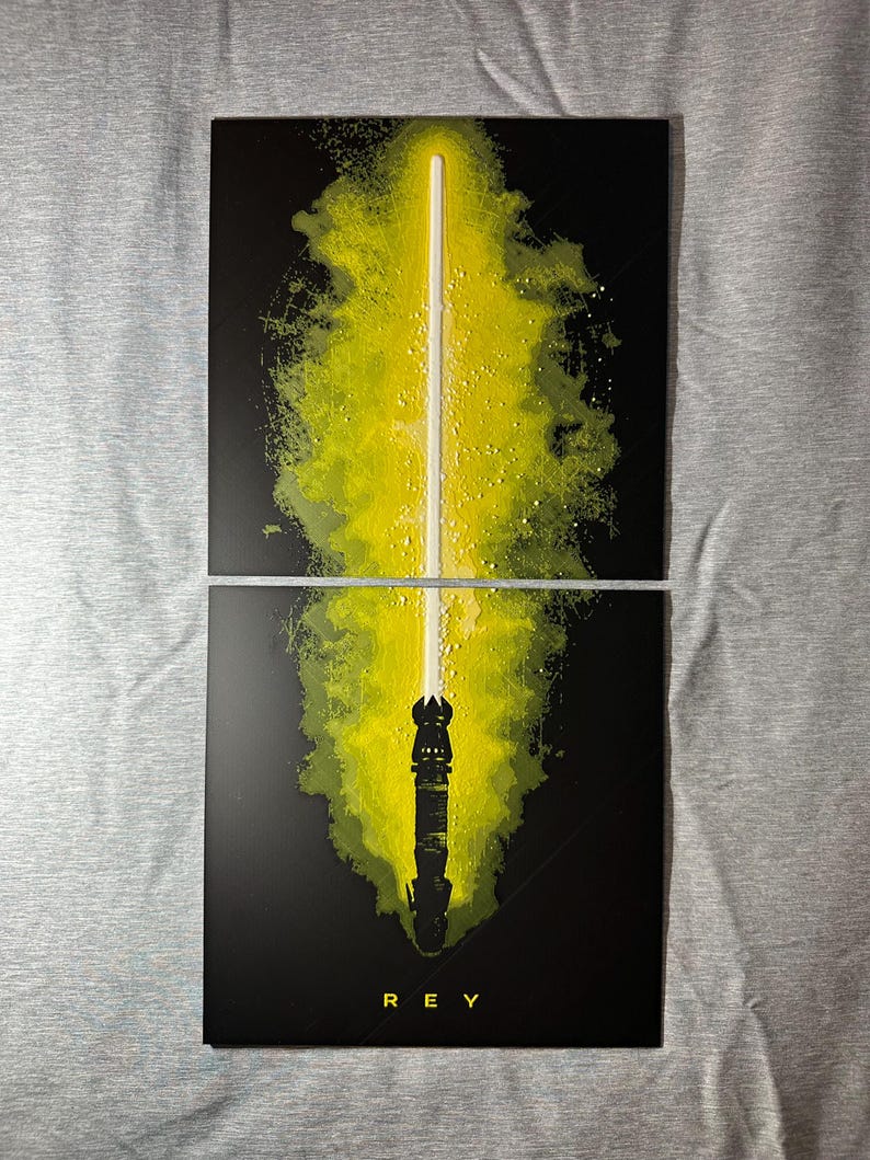K&ouml;nnte beinhalten: Zweiteiliges Kunstwerk mit einem gelben Lichtschwert vor schwarzem Hintergrund. Die Klinge des Lichtschwerts ist wei&szlig;, mit einem gelben und gr&uuml;nen Spritzeffekt. Das Wort "REY" ist unten in Wei&szlig; gedruckt.
