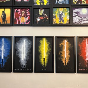 K&ouml;nnte beinhalten: F&uuml;nf gerahmte Kunstdrucke mit stilisierten Lichtschwertern in Blau, Wei&szlig;, Gelb, Orange und Rot. Jeder Druck hat einen schwarzen Rahmen und einen dunklen Hintergrund. Die Namen "Anakin", "Ahsoka Tano", "Rey", "Baylan Skoll" und "Darth Vader" sind unten aufgedruckt.