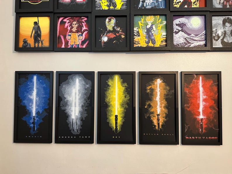 K&ouml;nnte beinhalten: F&uuml;nf gerahmte Kunstdrucke mit stilisierten Lichtschwertern in Blau, Wei&szlig;, Gelb, Orange und Rot. Jeder Druck hat einen schwarzen Rahmen und einen dunklen Hintergrund. Die Namen "Anakin", "Ahsoka Tano", "Rey", "Baylan Skoll" und "Darth Vader" sind unten aufgedruckt.