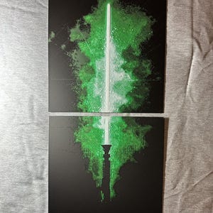 K&ouml;nnte beinhalten: Zweiteiliger Kunstdruck mit einem gr&uuml;nen Lichtschwert-Design auf schwarzem Hintergrund. Die Lichtschwertklinge ist wei&szlig;, mit einem gr&uuml;nen, strukturierten Effekt. Das Wort "LUKE" ist unten gr&uuml;n gedruckt.