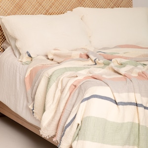 May include: A bed with white bedding and two white pillows. A striped blanket in pastel colors of pink, green, blue, and gray is draped over the bed. The headboard is made of woven wood.