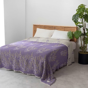 Pode incluir: Uma manta de tecido waffle roxo e creme com um padrão floral de lavanda. A manta está colocada sobre uma cama com uma cabeceira de madeira.