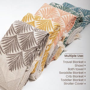 May include: A stack of five Turkish towels in various colors, including gray, yellow, peach, and green. The towels feature a geometric pattern of stylized palm leaves. The text "Multiple Use: Travel Blanket. Shawl. Bath towel. Swaddle Blanket. Crib Blanket. Toddler Blanket. Stroller Cover." is printed below the towels.