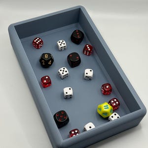 May include: A gray rectangular dice tray with a variety of dice inside. The dice are red, black, white, and yellow. The tray is made of a smooth, hard plastic.