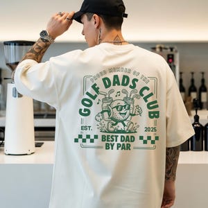 May include: Cream-colored t-shirt with a green graphic on the back that reads "Golf Dads Club." The graphic features a golf ball character with sunglasses and golf clubs. The text "Best Dad by Par" is also included.
